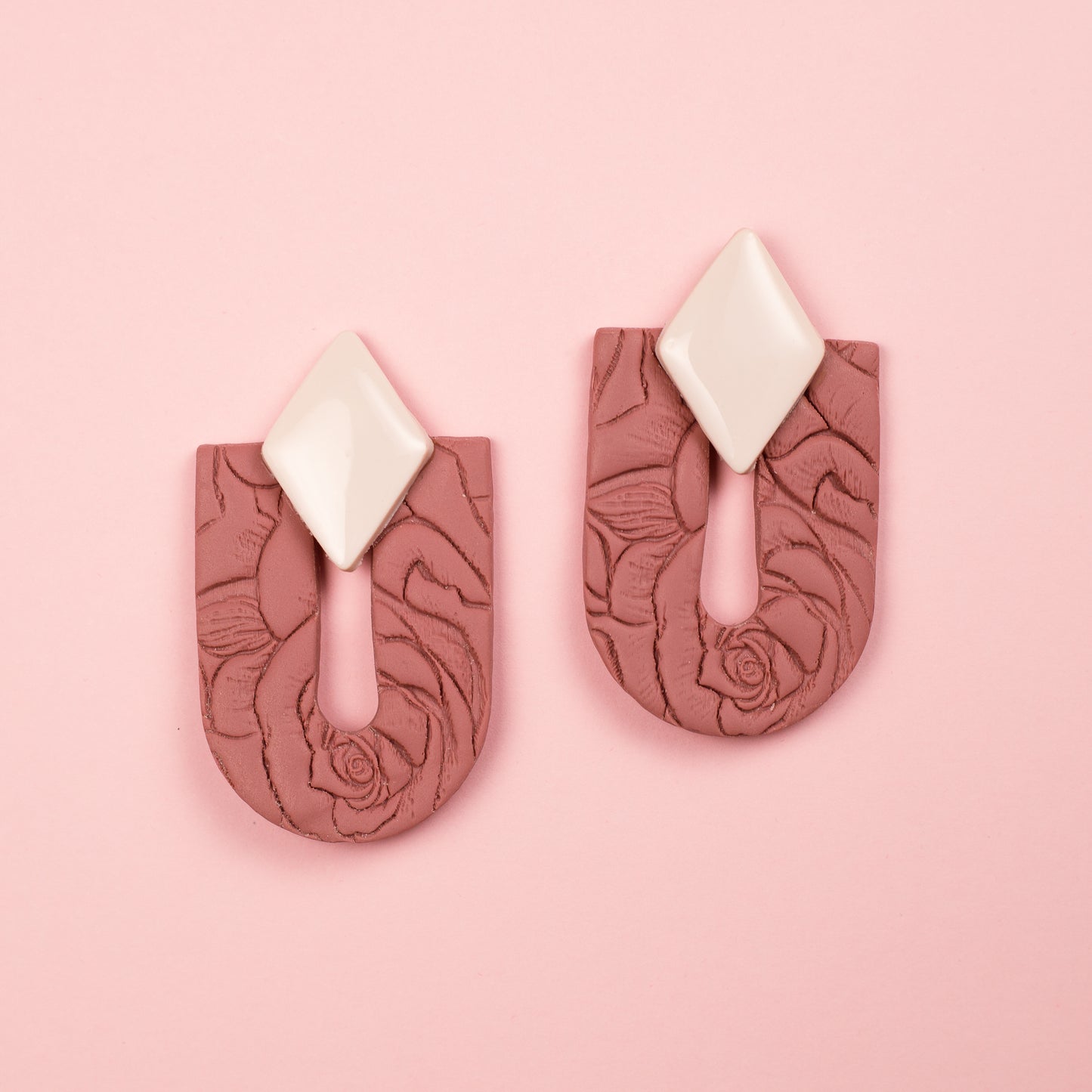 Pendientes hechos a mano de arcilla polimérica y acero inoxidable con forma de arco y rombo y textura floral en tonos rosas y blancos.